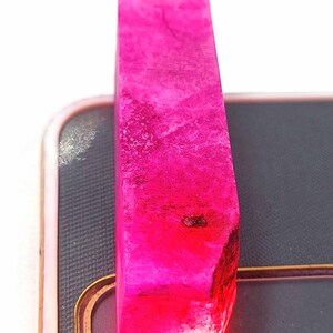 Peut inclure: Une pierre précieuse rectangulaire de couleur rose vif, taillée grossièrement, avec un léger éclat. La pierre précieuse est posée sur une surface noire.