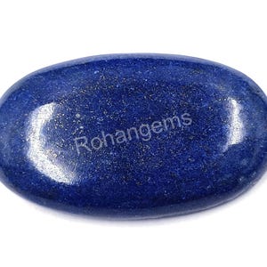 Pode incluir: Gema ovalada de lápis-lazúli polido. A pedra azul escura tem manchas douradas e o texto "Rohangems" impresso na superfície. A gema está sobre um fundo branco.