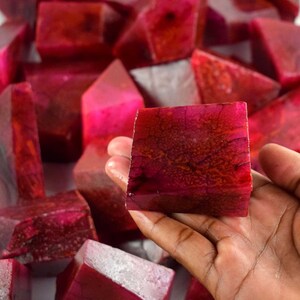 May include: A close-up of a hand holding a deep red, square-shaped, polished gemstone. The gemstone has a marbled, textured surface and is surrounded by other similar gemstones.