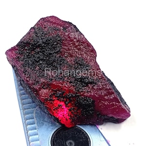 May include: A raw ruby gemstone, displaying a deep red colour with a dark, textured surface. The gemstone is irregularly shaped and appears unpolished. The text "Rohangems" is visible on the surface.