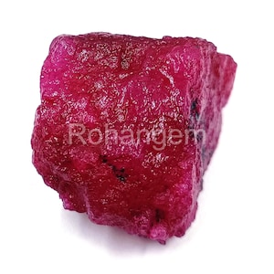 May include: A rough-cut, raw ruby gemstone with a deep red hue. The surface has a textured appearance, and the stone is irregularly shaped. The text "Rohangem" is visible on the surface of the stone.