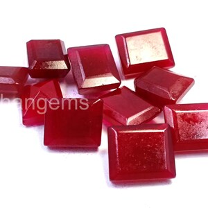 May include: A collection of faceted, square-cut, deep red gemstones. The gems are scattered across a white surface, showcasing their polished surfaces and varying shapes. The image highlights the rich color and clarity of the stones.