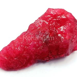 Può includere: Una gemma rosso vivo grezza con una superficie strutturata. La pietra ha una forma naturale e irregolare ed è esposta su uno sfondo bianco. Il testo "Rohangem" è visibile sulla superficie della pietra.