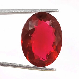 Op de afbeelding: Een dieprode ovale edelsteen met een facetgeslepen vorm. De edelsteen wordt vastgehouden door een pincet.