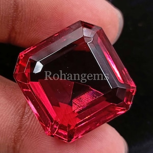 Puede incluir: Una piedra preciosa roja oscura con un corte cuadrado esmeralda. La piedra preciosa está facetada y tiene un alto pulido.