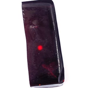 Puede incluir: Objeto rectangular burdeos oscuro con una parte superior redondeada y un punto rojo. El objeto tiene un acabado brillante y parece estar hecho de un material sólido. Hay arañazos e imperfecciones en la superficie.