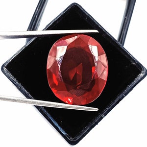 May include: A deep red oval gemstone with a faceted cut, displayed in a black velvet box.