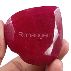 Puede incluir: Una gema facetada de color rojo intenso con el texto "Rohangem" impreso en la superficie. La gema tiene una superficie pulida y reflectante y se sostiene en una mano. La gema tiene una forma irregular única.