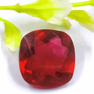 May include: A deep red, faceted gemstone with a cushion cut. The gemstone is clear and has a slight pink hue.