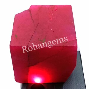 Pode incluir: Uma grande gema de rubi vermelho bruto com um acabamento natural não polido. O rubi é iluminado por baixo, mostrando sua cor vermelha intensa. O texto "Rohangems" é visível na superfície do rubi.
