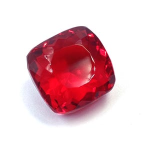 May include: A vibrant, cushion-cut gemstone with a deep, saturated red color. The stone features a faceted surface that reflects light, creating a brilliant, sparkling appearance across its square-like shape with rounded corners and edges.