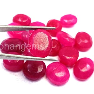 May include: A collection of polished, oval-shaped gemstones in varying shades of pink and red. One gem is held by tweezers, showcasing its smooth surface. The image includes the text "ohangems".