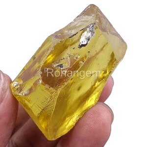 Puede incluir: Una gema amarilla translúcida en bruto con una estructura cristalina. La piedra tiene una forma natural e irregular y una superficie pulida y reflectante. El texto "Rohangem" es visible.