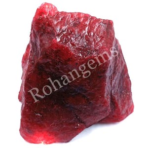 Puede incluir: Una gran gema roja rugosa con una veta rojo oscuro, casi negra, que la atraviesa. La gema es un rubí en bruto sin pulir.
