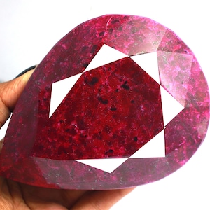 May include: A large, pear-shaped ruby gemstone with a faceted cut. The ruby is a deep red color with a slightly pink hue. The gemstone is set in a clear, faceted setting.