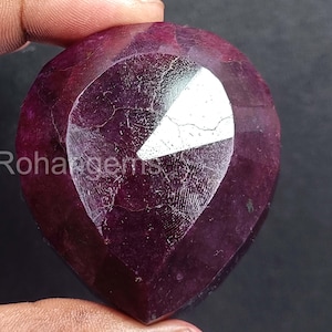 Puede incluir: Una gema de rubí facetada en forma de corazón. La gema de color rojo intenso tiene una superficie pulida con inclusiones internas visibles y una gran faceta reflectante. El texto "Rohangems" es visible.