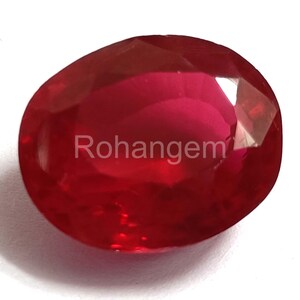 Puede incluir: Una gema de rubí facetada de forma ovalada. La gema es de color rojo intenso y saturado con una superficie pulida. El texto "Rohangem" es visible en la superficie del rubí.