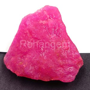 Puede incluir: Una gema de rubí en bruto y sin cortar, de un tono rosa vibrante. La superficie tiene un aspecto texturizado, con la palabra "Rohangeu" impresa. La gema se muestra sobre un fondo blanco y negro.