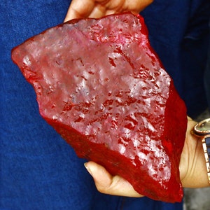 May include: A large, rough, red gemstone. The gemstone is being held in a hand.