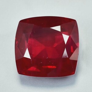 May include: A faceted, cushion-cut ruby gemstone. The gem is a deep, saturated red colour with sharp, defined facets that reflect light. The shape is square with rounded corners, and the overall appearance is polished and brilliant.