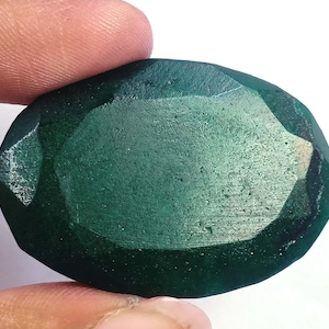 Puede incluir: Una gema verde oscuro, facetada y de forma ovalada. La superficie pulida de la gema refleja la luz. La piedra se sostiene entre dos dedos, destacando su tamaño y color. La gema parece ser una esmeralda natural.