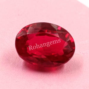 Puede incluir: Una piedra preciosa ovalada de color rojo intenso con un corte facetado. La piedra preciosa está sobre una superficie rosa y el texto "Rohangems" es visible en el centro de la piedra.