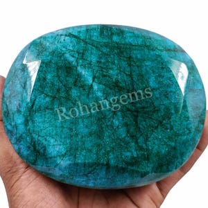 Puede incluir: Una gran gema de esmeralda de forma ovalada, de color verde intenso y con inclusiones visibles. La gema se sostiene en una mano, y el texto "Rohangems" es visible en el fondo.