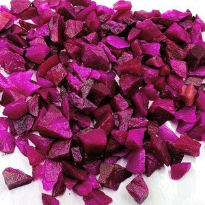 May include: A large pile of small, irregularly shaped, magenta-colored glass pieces. The pieces are scattered on a white surface.
