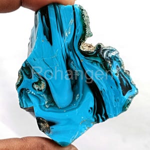 May include: A vibrant, irregularly shaped gemstone with a striking blend of turquoise and black hues. The polished surface showcases a swirling pattern, with areas of rough, textured edges. The gemstone is held by a hand.