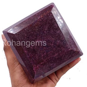以下が含まれることがあります： 深いマゼンタ色の大きな正方形にカットされた宝石。 ファセット加工されたエッジが光を捉え、石の内部の質感を明らかにします。 「Rohangems」の文字が石の側面に表示されています。