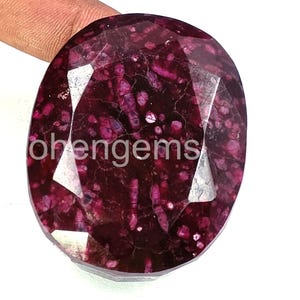 Puede incluir: Una gema grande y facetada de color burdeos intenso con patrones internos de rosa y blanco más claros. La piedra de forma ovalada tiene una superficie pulida y el texto "ohengems" es visible.