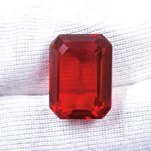 May include: A faceted, rectangular-cut, red gemstone. The gem has a vibrant, transparent appearance and is set against a white, textured background. The gemstone's edges are precisely cut, reflecting light.
