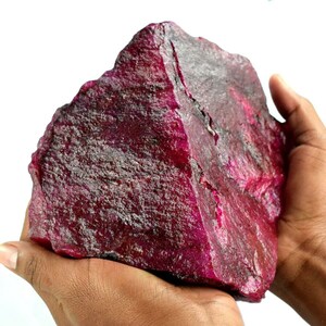May include: A large, rough-hewn ruby gemstone with a deep red color. The raw mineral is held in two hands, showcasing its irregular shape and textured surface. The ruby appears to be uncut and in its natural state.