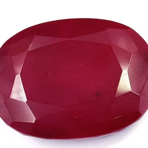 Puede incluir: Una gema de rubí facetada de forma ovalada. La gema es de un color rojo intenso y rico, con múltiples facetas que capturan la luz. La gema se encuentra sobre un fondo blanco, resaltando su color y talla.
