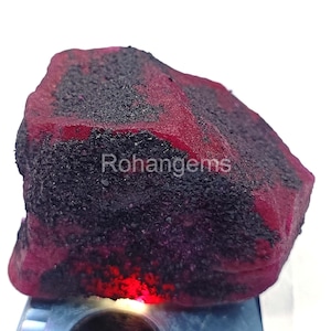 May include: A raw ruby gemstone, displaying a rich red colour with dark, granular inclusions. The stone's surface has a textured appearance, and the word "Rohangems" is visible on the surface.
