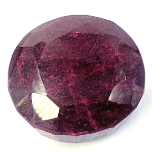Puede incluir: Una gema redonda y facetada de color burdeos intenso. La gema tiene múltiples facetas que reflejan la luz, creando un efecto brillante. La piedra está sobre un fondo blanco.