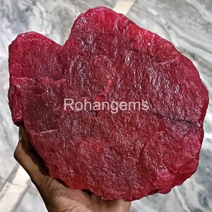May include: A large, rough, red ruby gemstone. The ruby is a deep, rich red color and has a natural, unpolished surface. The ruby is being held in a hand.