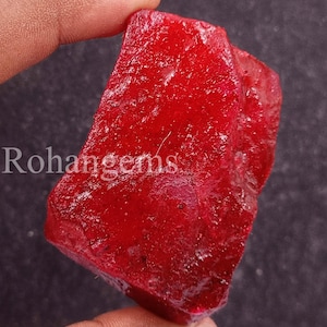 May include: A rough-cut, deep red gemstone with a textured surface. The gemstone is held between fingers, and the text "Rohangems" is visible on the side. The stone has a raw, natural appearance.