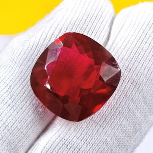 May include: A faceted, cushion-cut gemstone in a vibrant red hue. The gem is held against a white fabric background, with a yellow backdrop. The gemstone's cut creates multiple reflective surfaces.