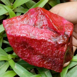 May include: A large, rough, red gemstone with a deep, rich color. The gemstone has a natural, unpolished surface.