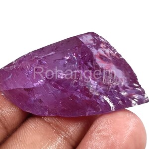 Può includere: Una gemma di ametista naturale grezza in tonalità viola. La pietra ha una struttura cristallina ed è tenuta in mano. La parola "Rohangem" è visibile sulla pietra.