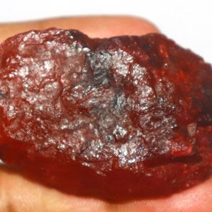 May include: A rough, uncut gemstone with a deep red hue. The surface of the stone has a textured appearance, with visible crystalline structures and variations in color. The gemstone is held in a hand, showcasing its natural form.