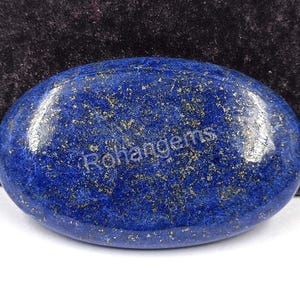 Può includere: Una gemma di lapislazzuli lucidata, di forma ovale. La pietra blu scuro è punteggiata da inclusioni dorate di pirite, creando un effetto scintillante. La superficie è liscia e riflettente, con il testo "Rohangems" visibile.