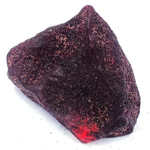 Puede incluir: Una piedra de color púrpura oscuro, de forma irregular, con una superficie rugosa y texturizada. La piedra tiene un color profundo, casi negro, con toques de rojo y una textura moteada y arenosa. El borde inferior tiene un tono rojo brillante.