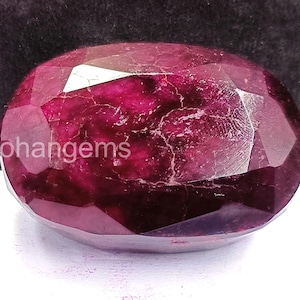 May include: A faceted, oval-shaped ruby gemstone with a deep red hue. The gem has visible internal inclusions and a polished surface. The text "Rohangems" is visible on the left side of the gem.