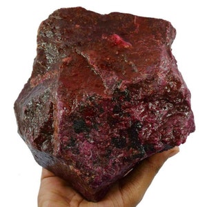 May include: A large, rough, red ruby gemstone. The ruby is a deep red color with a slightly purplish hue. It has a natural, unpolished finish.