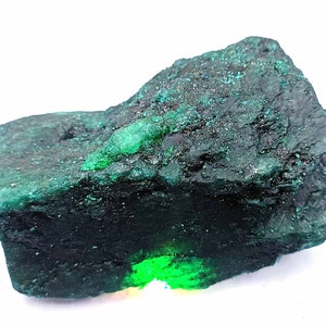 Può includere: Un grande cristallo di smeraldo verde scuro ruvido con una sezione liscia verde brillante. Il cristallo è su uno sfondo bianco.