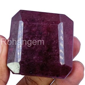May include: A large, faceted, rectangular gemstone with a deep burgundy color. The gem has a polished surface with multiple facets, reflecting light. The word "Rohangem" is visible on the side of the gem. The gem is held in a hand.