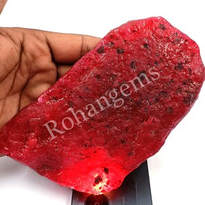 Puede incluir: Una gran gema de rubí rojo intenso con una superficie rugosa y natural. La gema se sostiene en una mano, y la luz brilla a través de ella, destacando su color rojo.