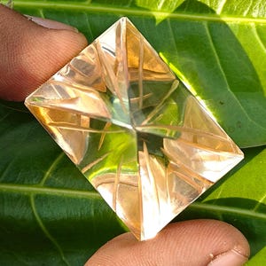 Könnte beinhalten: Ein facettierter, rechteckiger Edelstein mit warmem, goldenem Farbton. Der Stein weist komplizierte, lineare Schnitte auf, die ein geometrisches Muster erzeugen. Der Stein wird vor einem Hintergrund aus grünem Laub gehalten.